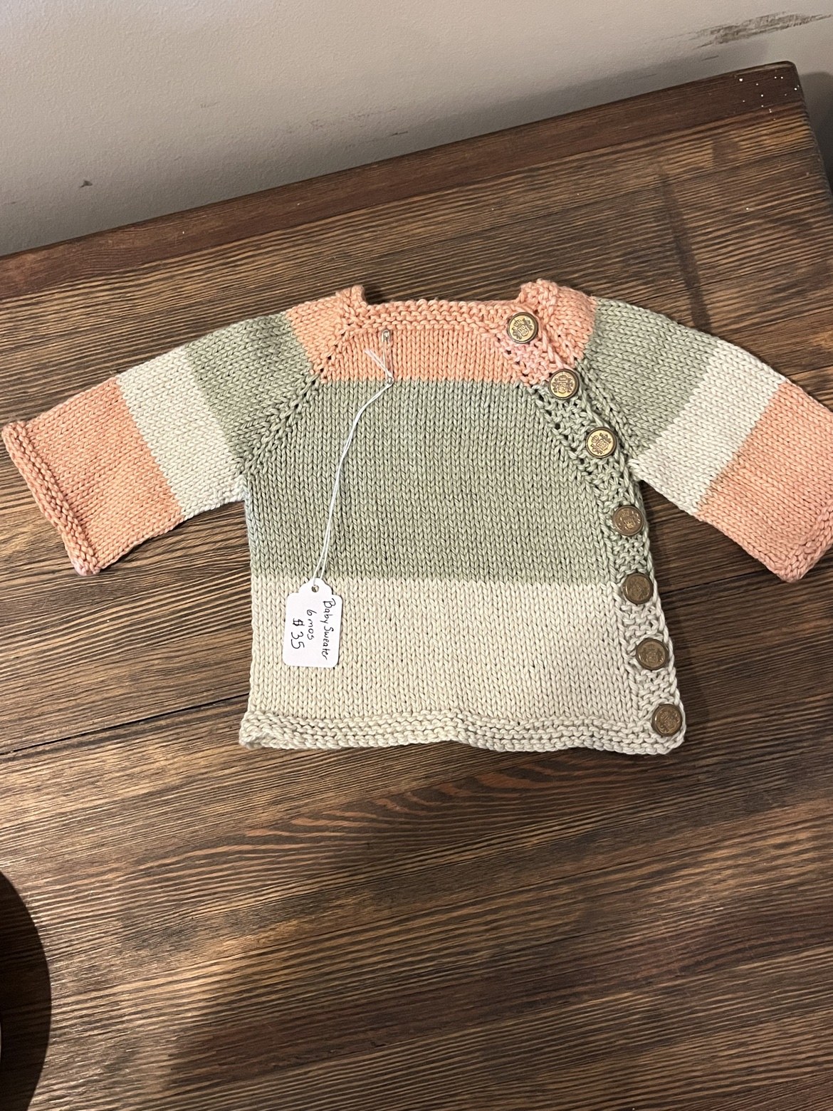 Full view of a hand-knit baby sweater in sage, cream, and peach with bronze-tone side buttons