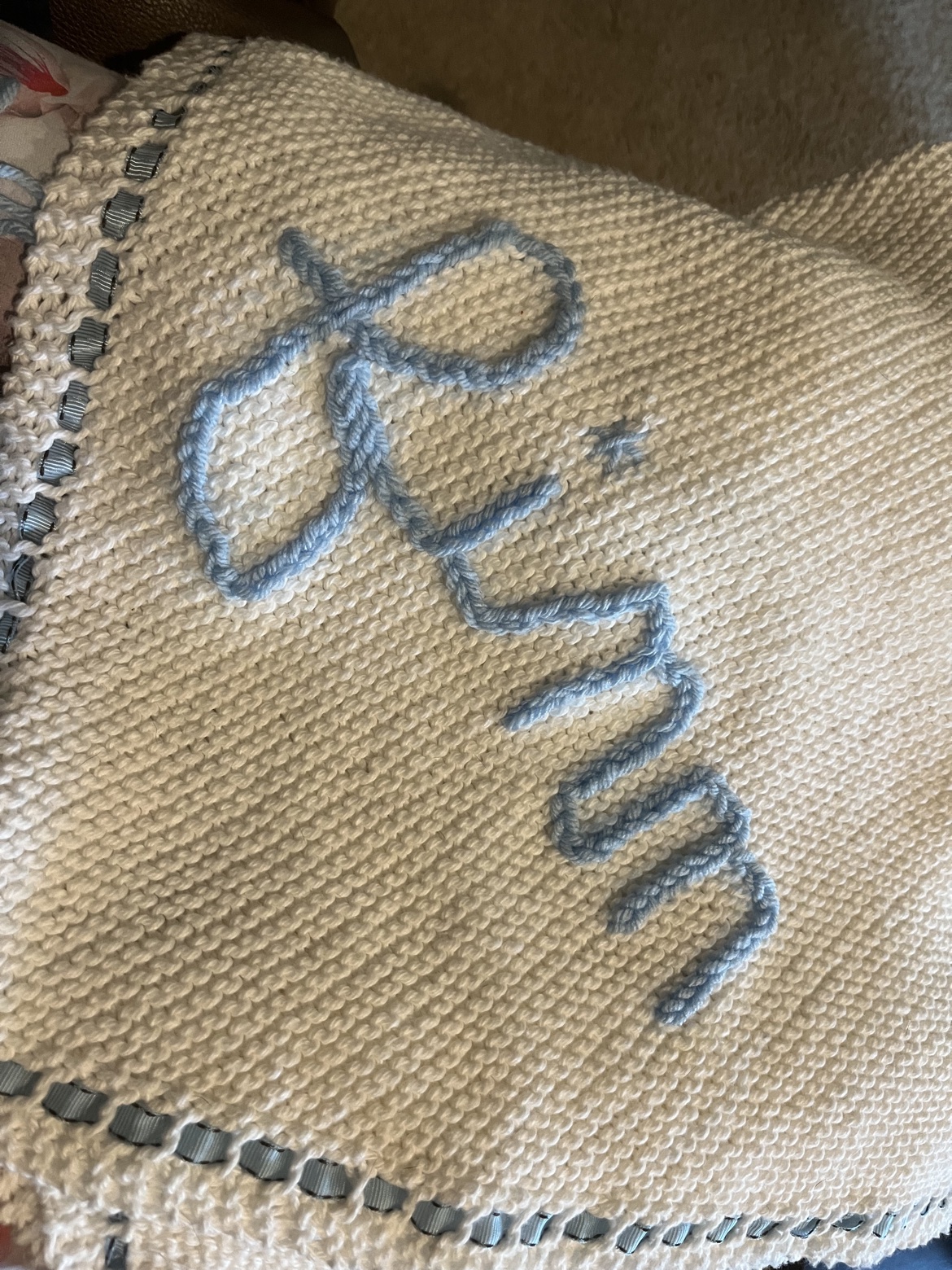 Cream hand-knit blanket with blue yarn lettering and ribbon trim