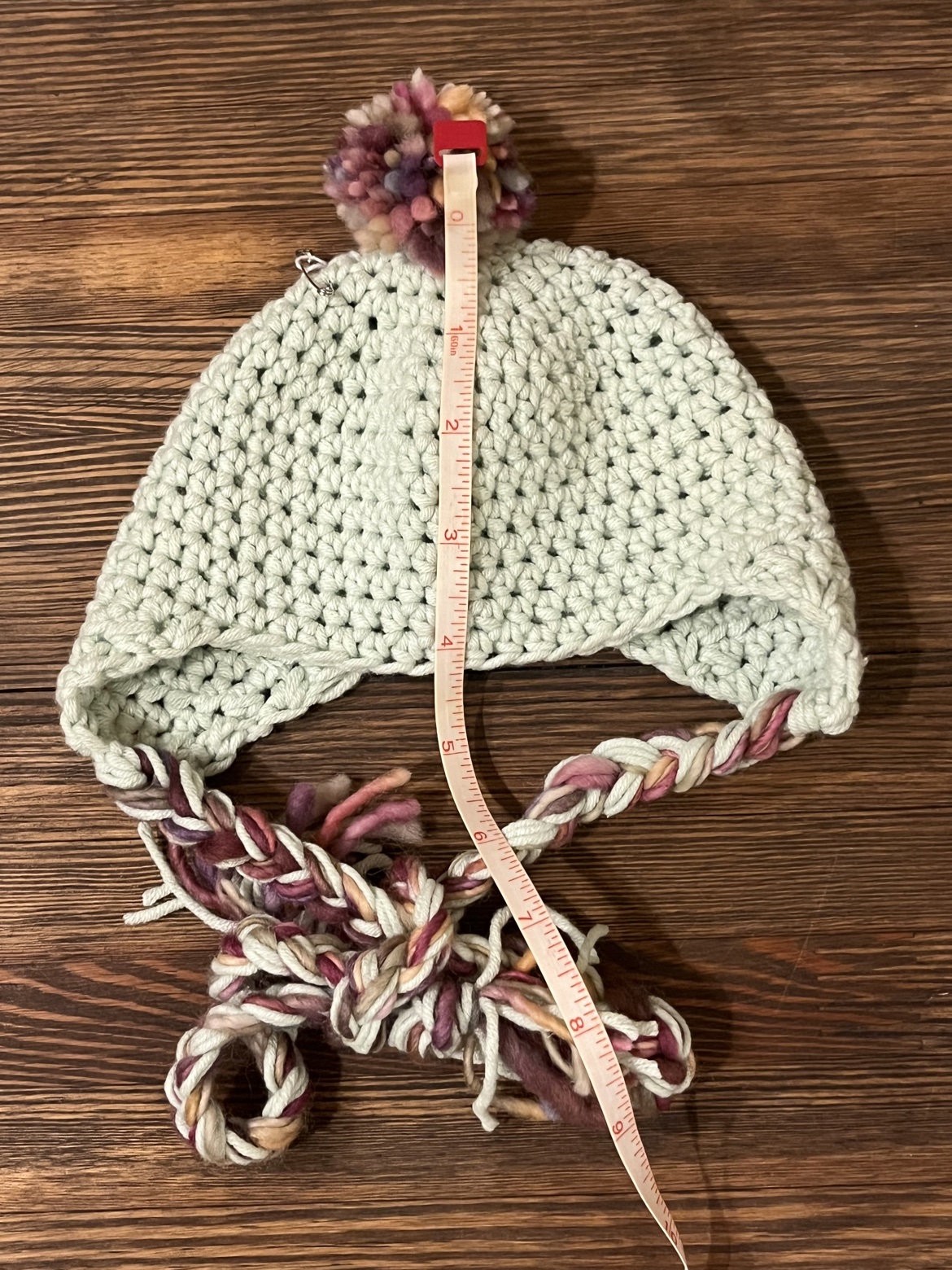 Baby ear flap hat shown with a measuring tape from pom-pom to ties